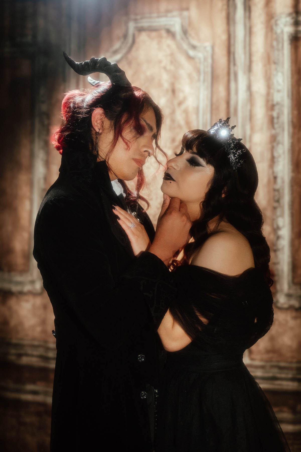 Couple in a horned headpiece and black crown face to face with a hand on a chin for dark romantic engagement photography