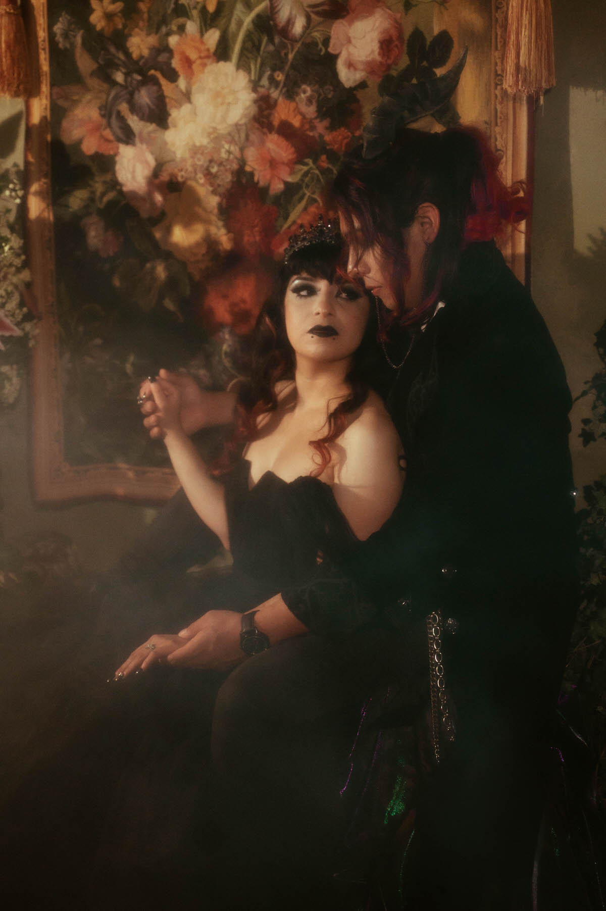 Seated couple holding hands in fog before a floral tapestry during a mood engagement session in Dallas