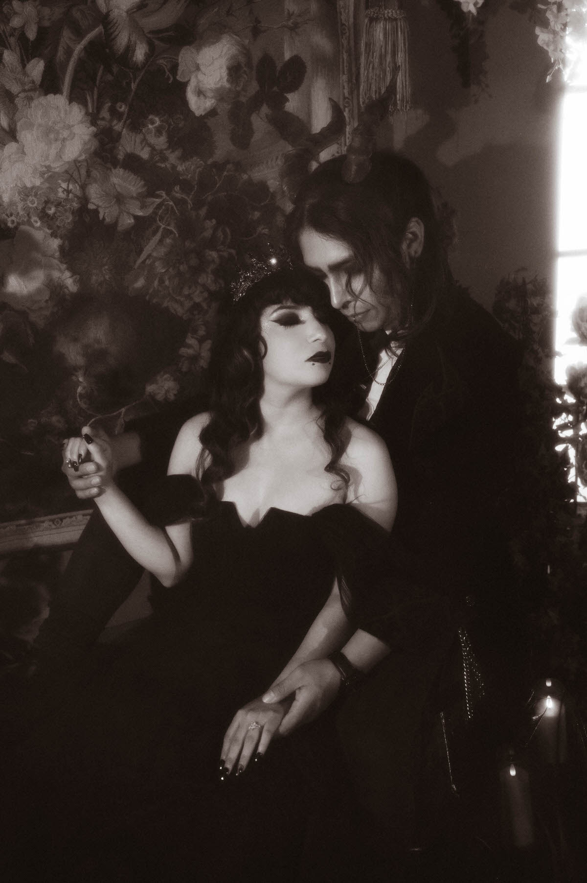 Black and white portrait of a seated couple holding hands before a floral tapestry for gothic couples photography