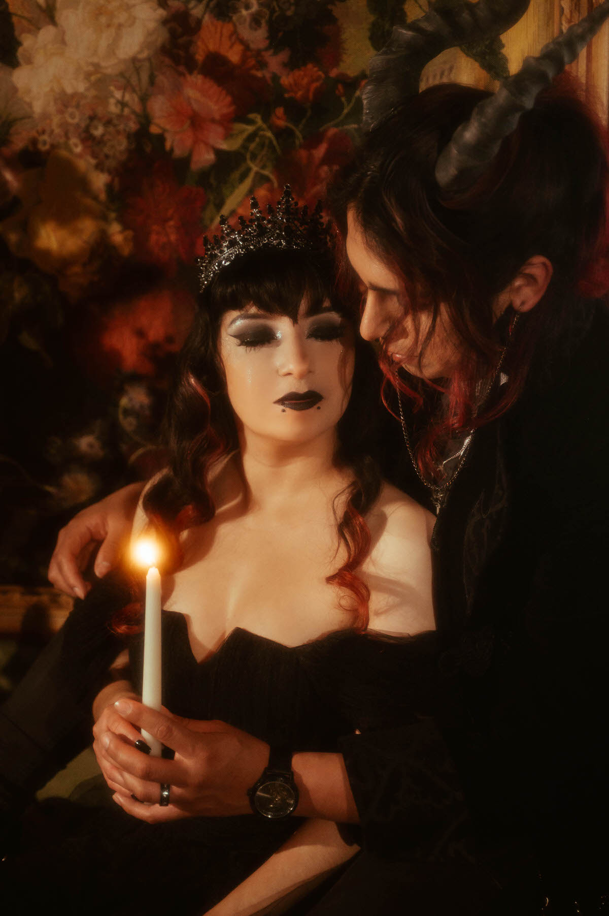 Couple sitting close holding a white taper candle before a floral tapestry for gothic bride and groom photos