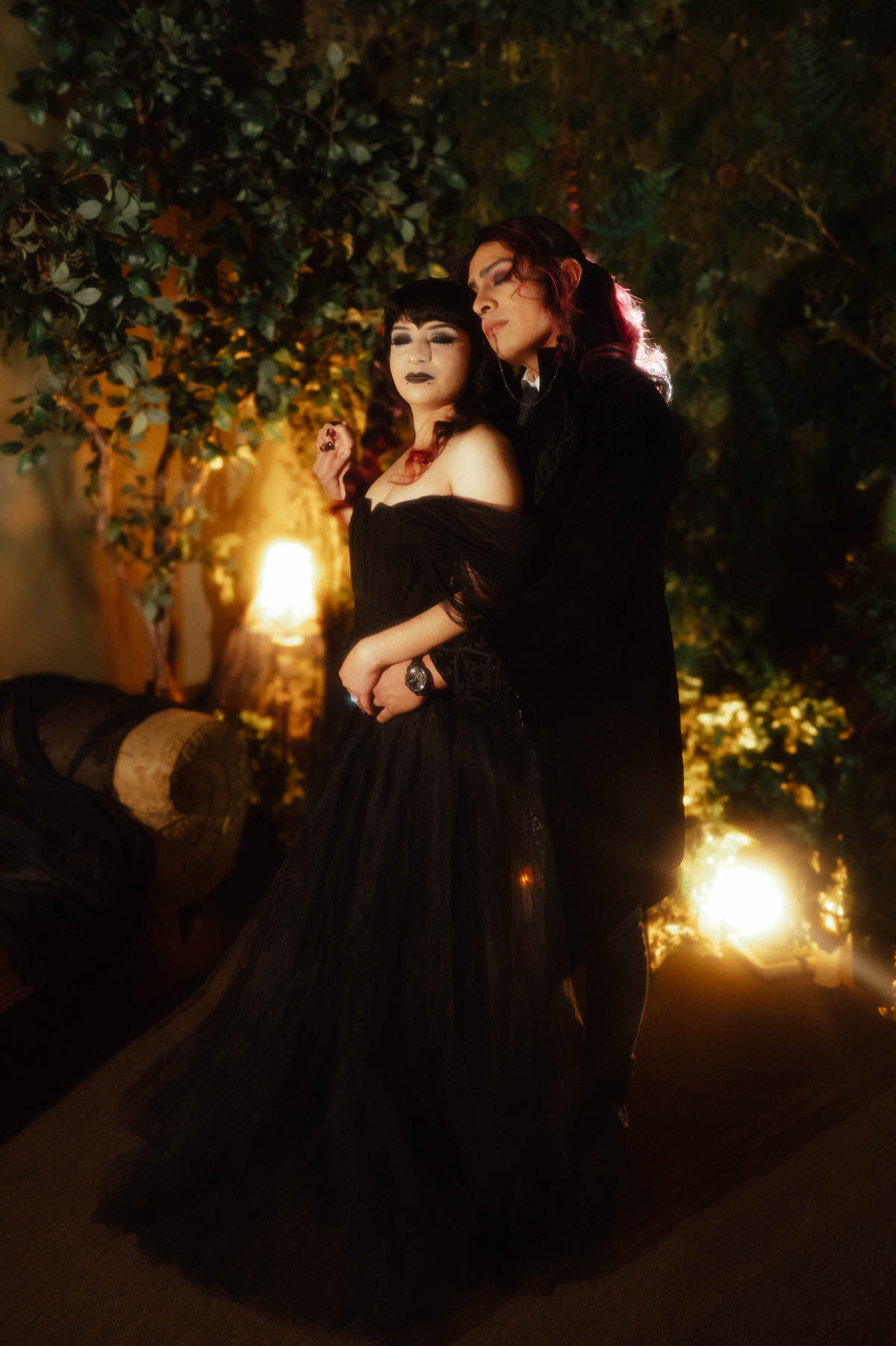 Couple in a black tulle gown and velvet coat embracing from behind in warm light for gothic bride and groom photos