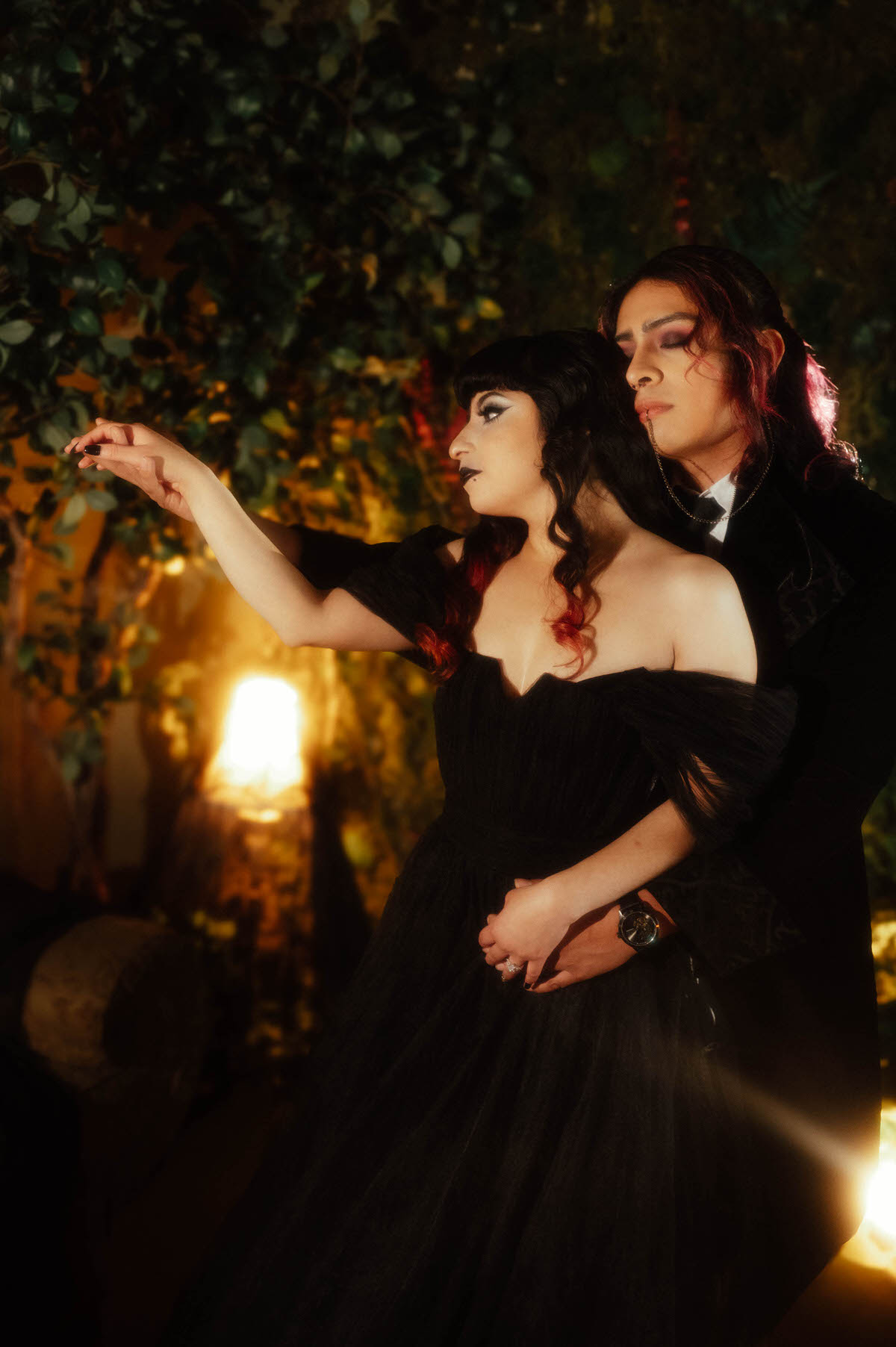 Couple embracing at the waist with one hand reaching toward leafy branches for dark romantic engagement photography