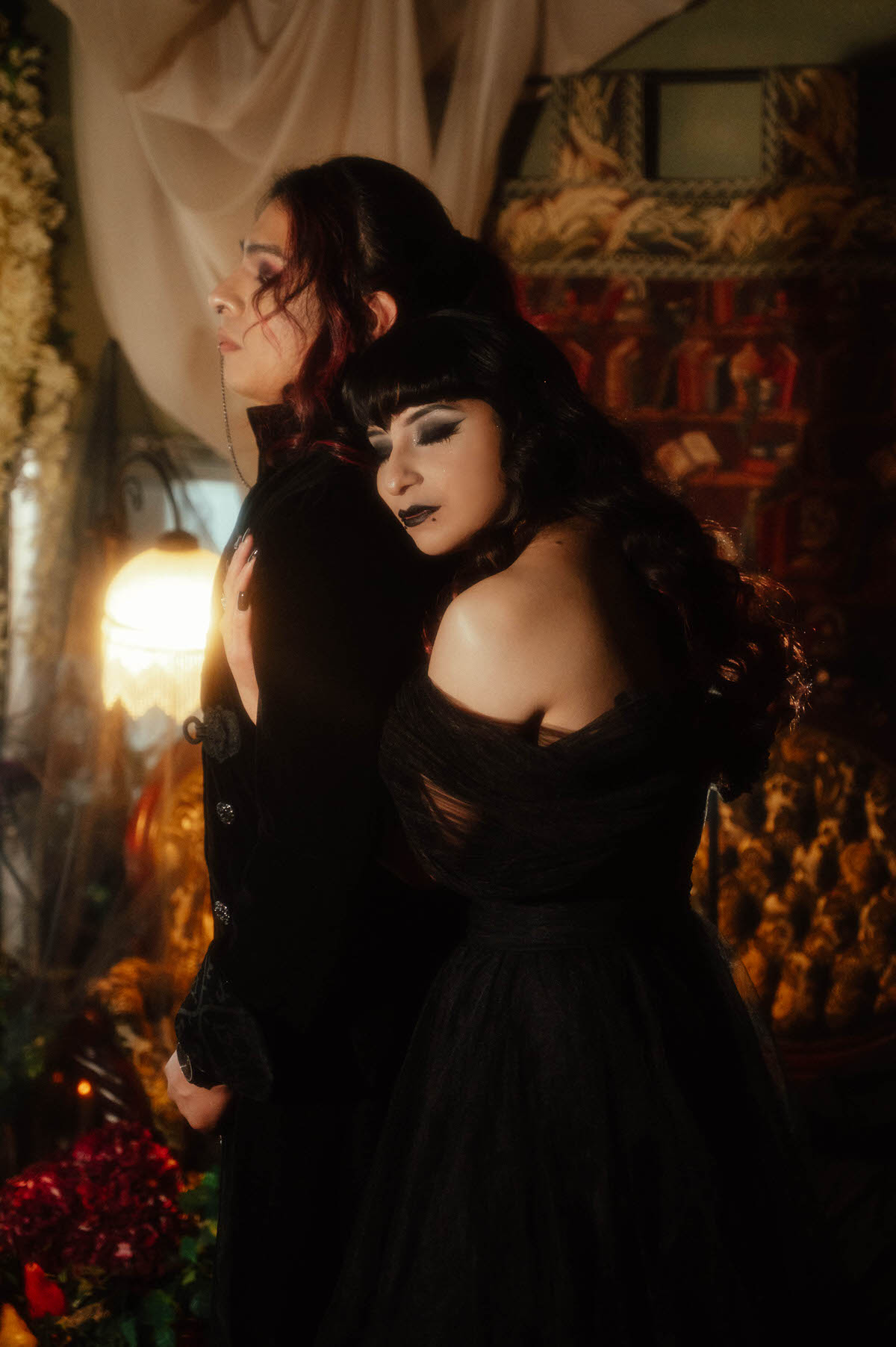 Couple in a black gown and black suit leaning together beside a glowing lamp for dark romantic engagement photography