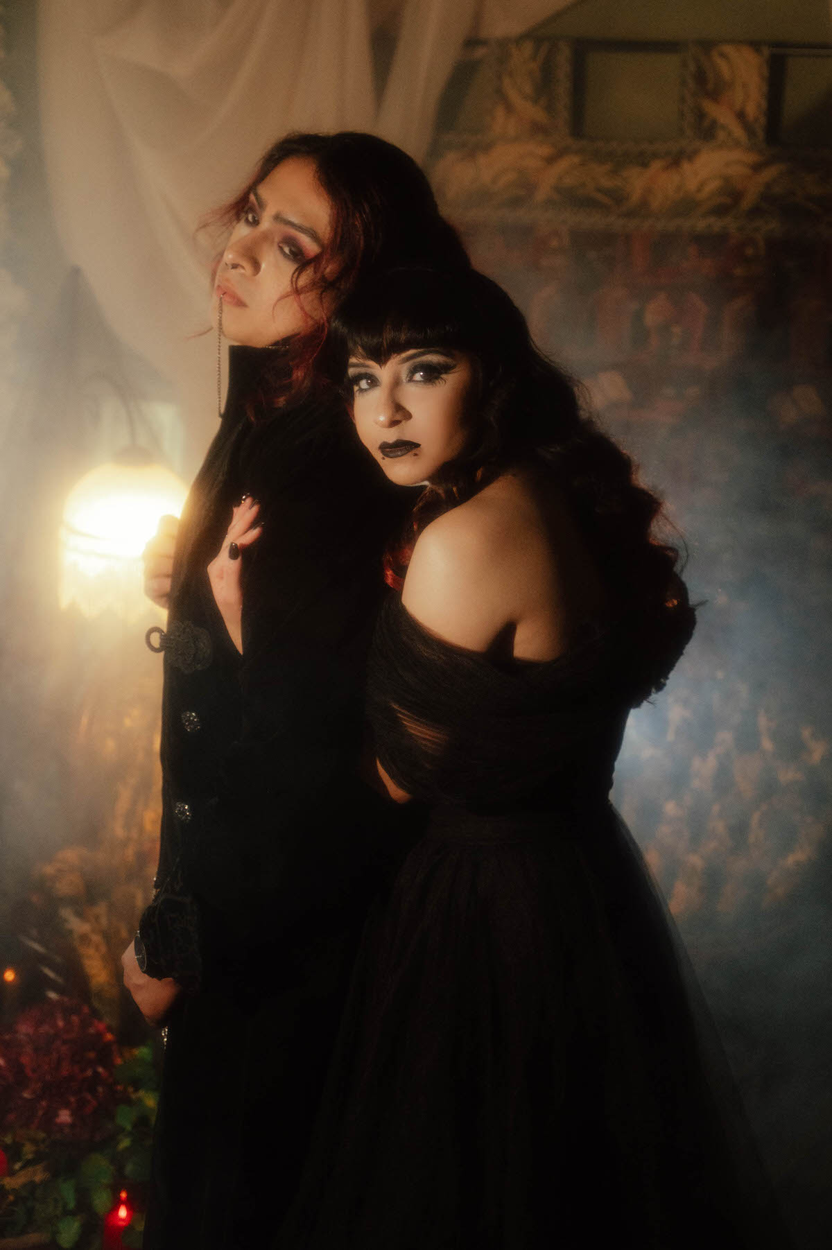Couple in a black gown and black suit looking at the camera beside a glowing lamp for gothic bride and groom photos
