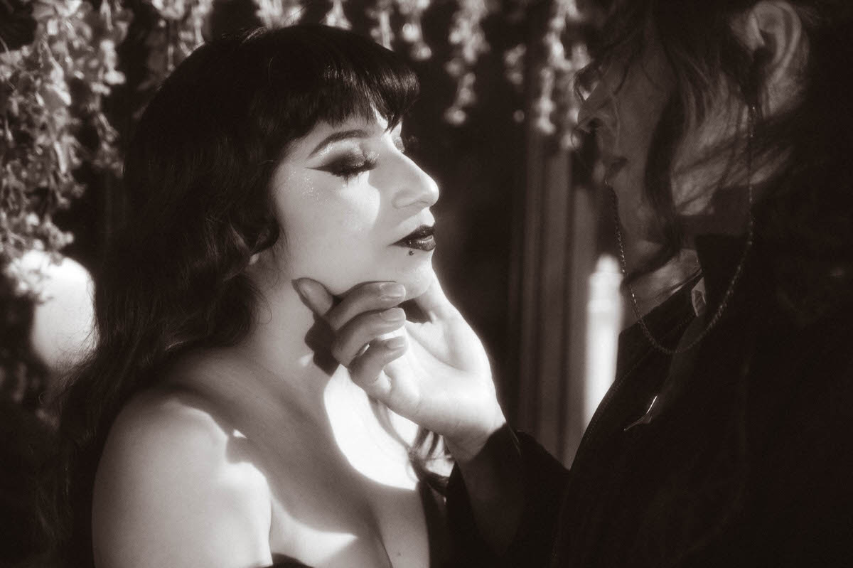 Black and white close-up of a hand cupping a partner's chin beside hanging flowers for gothic couples photography