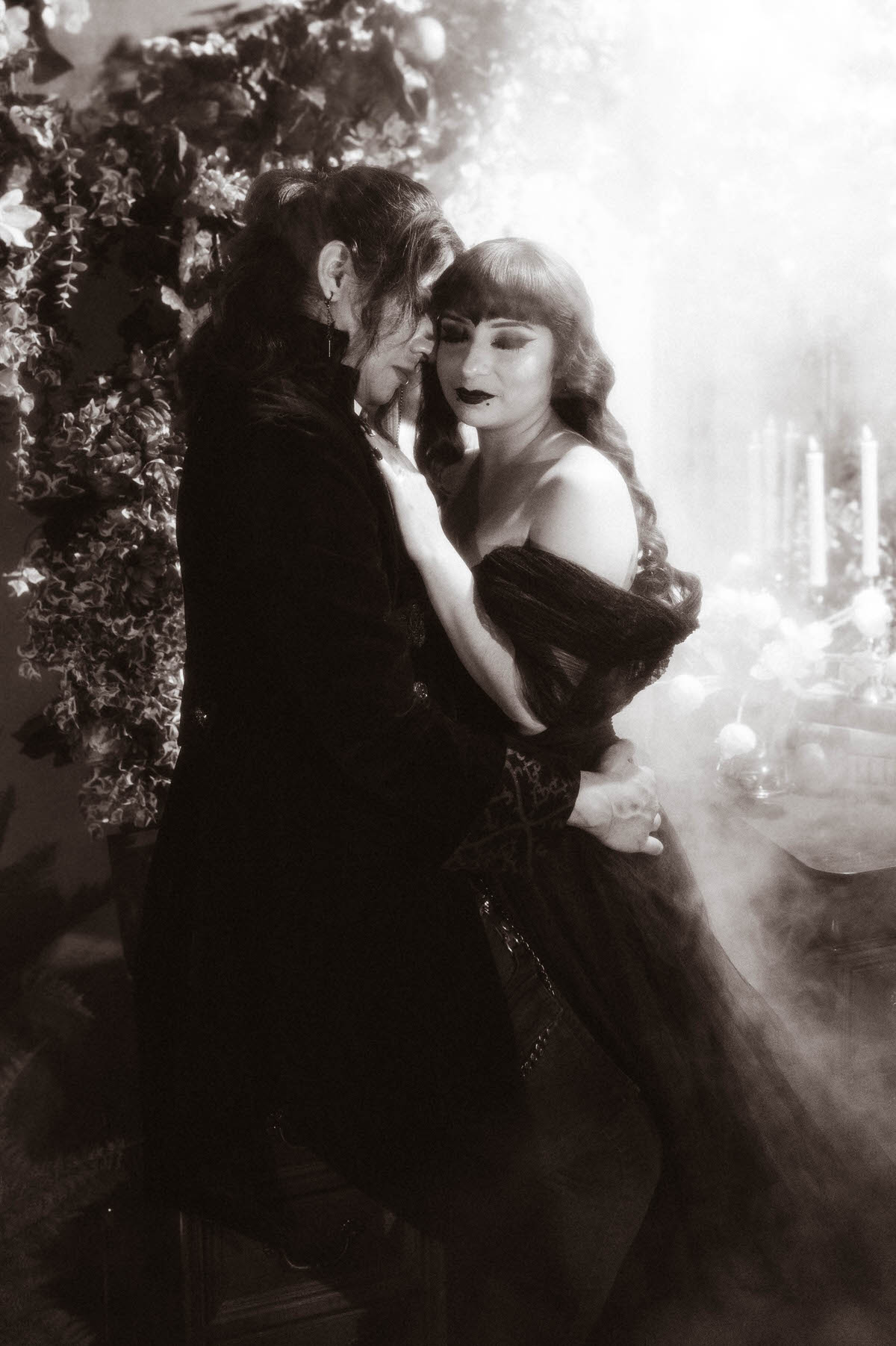 Black and white portrait of a couple embracing in fog near white candles and a leafy wall for gothic couples photography