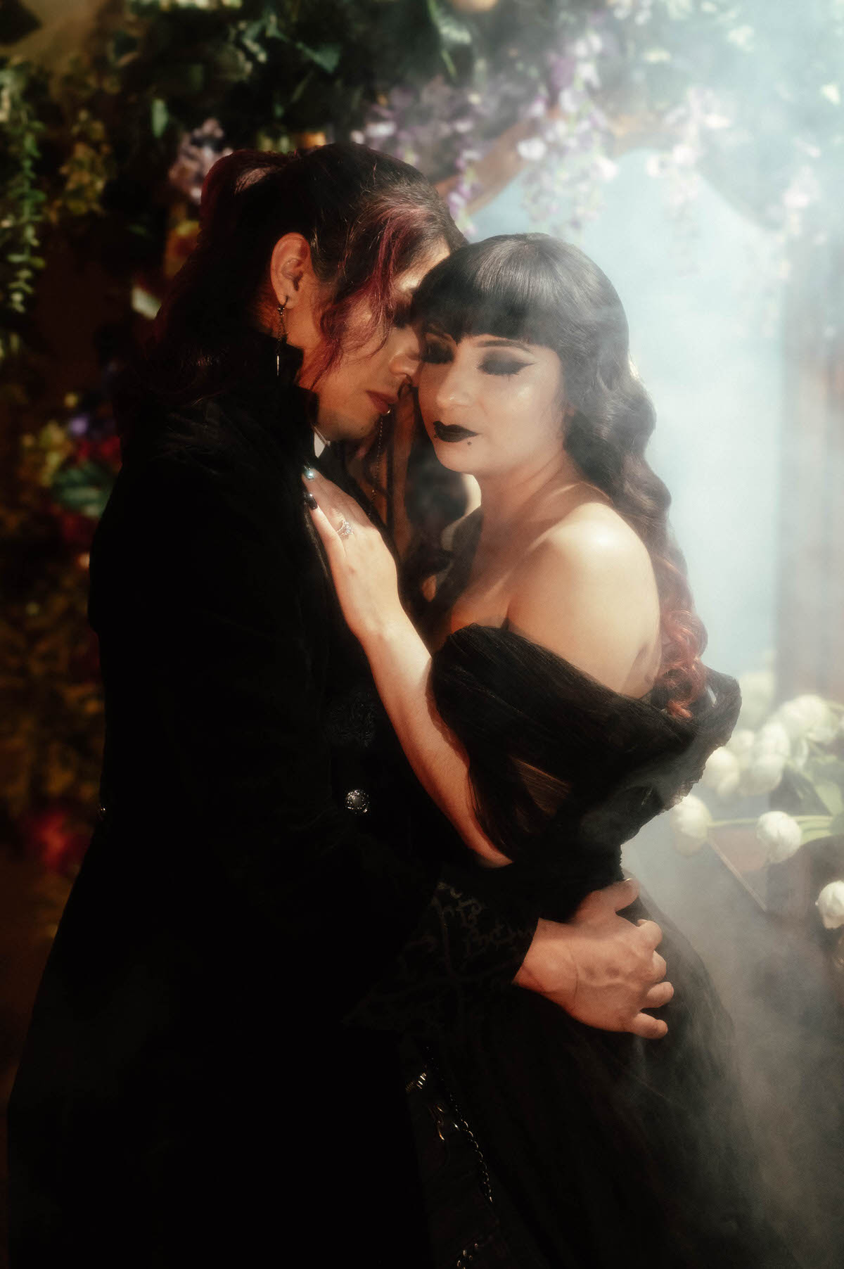 Couple embracing with foreheads together in fog near white flowers for gothic bride and groom photos