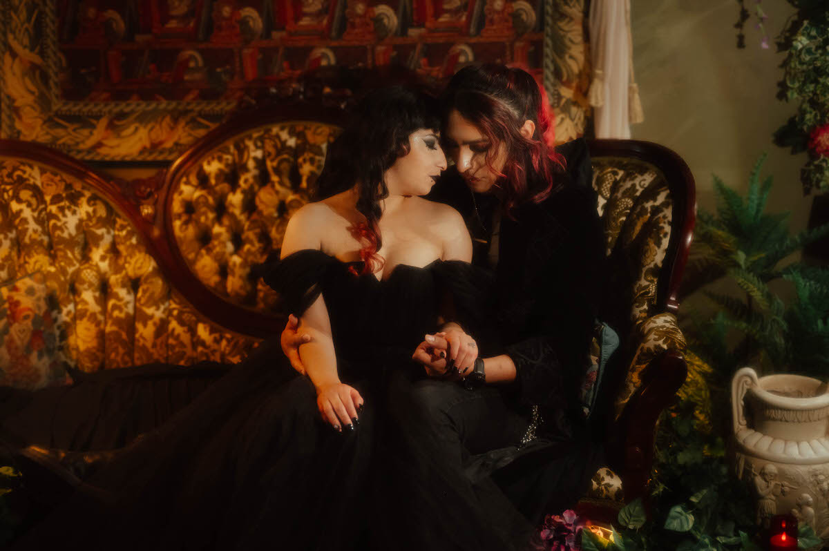 Couple in a black gown and black suit holding hands on a floral tufted sofa for dark romantic engagement photography