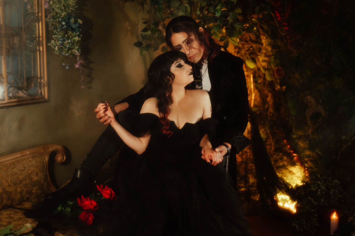 Couple holding hands on a chaise with red roses beside a leafy wall for a mood engagement session in Dallas