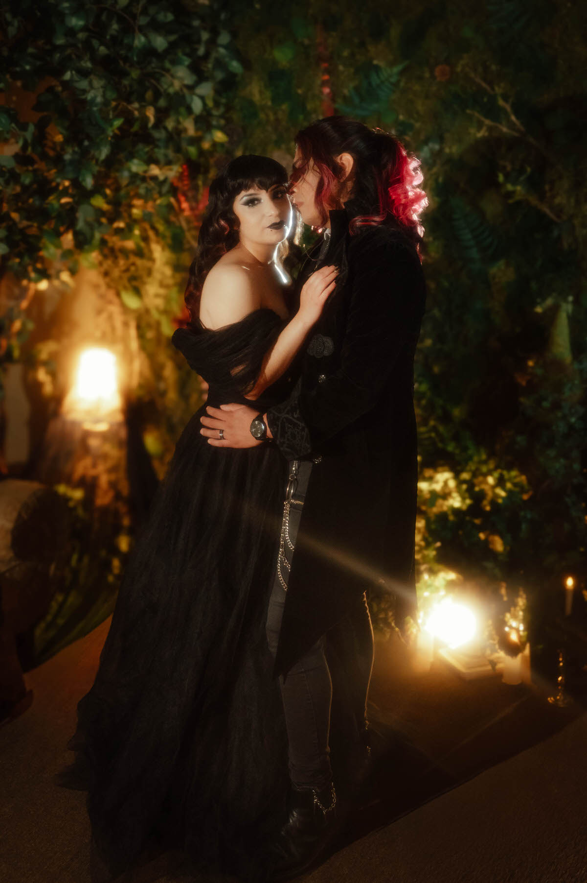 Couple embracing in front of a leafy wall with warm glowing lights during a mood engagement session in Dallas