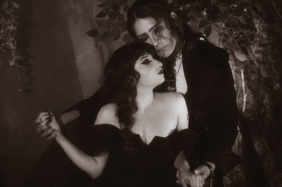 Black and white portrait of a seated couple holding hands before leafy branches for gothic couples photography