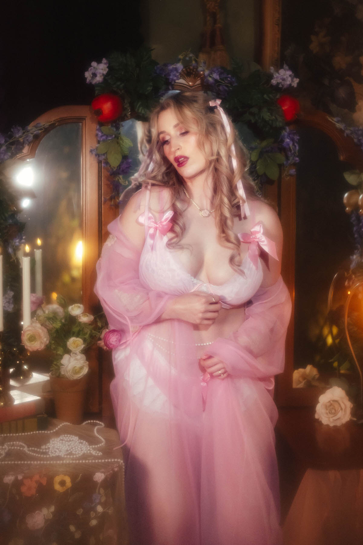 A woman in a vintage pink lingerie set and vintage sheer pink robe stands before a vintage vanity mirror crowned with pomegranates and purple flowers, hazy fog and candlelight glowing beside her, capturing the soft light intimate portraiture of this dreamy boudoir photoshoot with Royal Lune Photo.