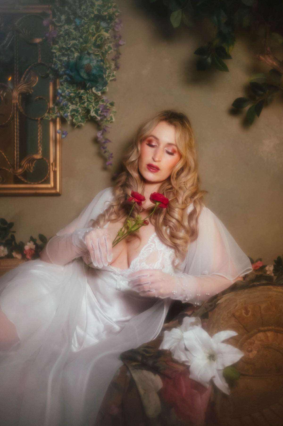A woman reclines on a gold tufted vintage sofa gazing down at dark pink florals in her pearl beaded gloved hand, wearing a vintage ivory satin slip dress and sheer chiffon robe near a gilded mirror and white lily blooms, capturing the whimsical boudoir portraiture Dallas style of Royal Lune Photo.