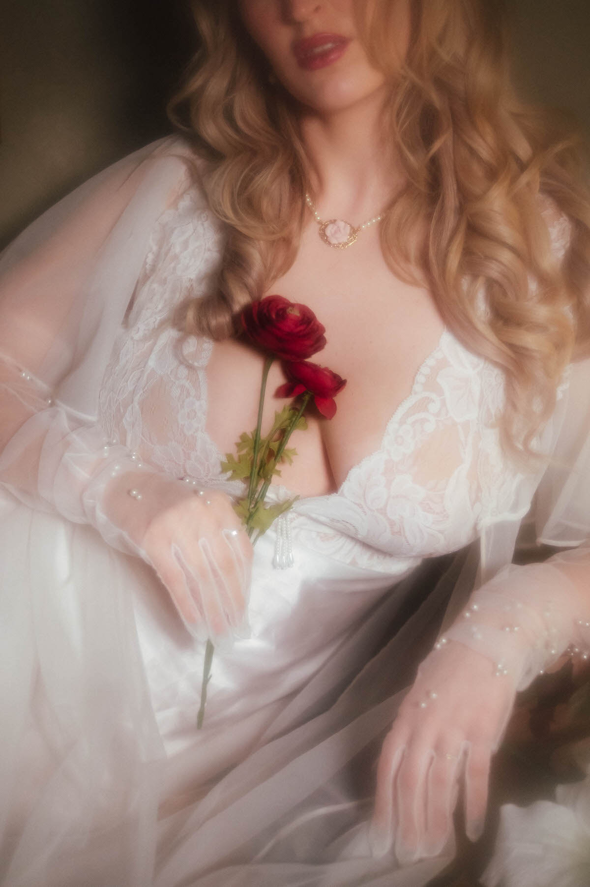 A close up shows dark pink florals held against a vintage ivory lace bodice and pearl necklace, with a pearl beaded glove resting nearby, highlighting the soft light intimate portraiture of this dreamy boudoir photoshoot with Royal Lune Photo.
