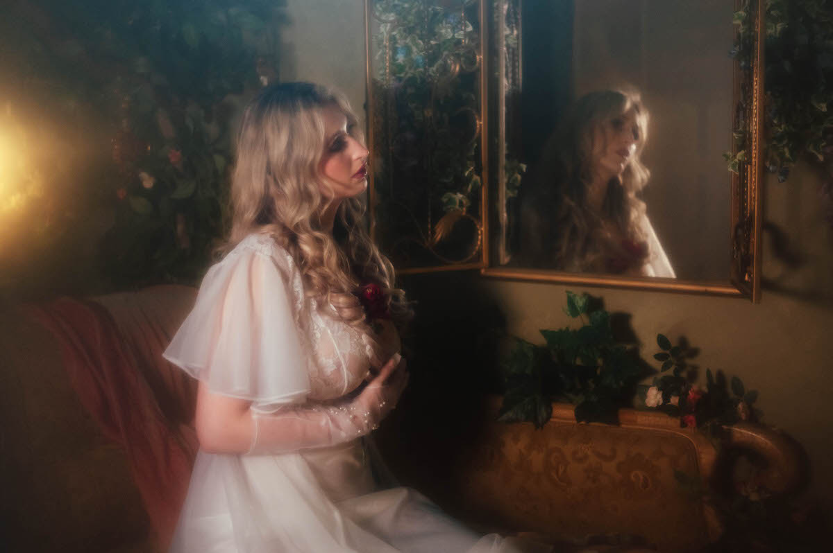 A woman kneels on a rose colored vintage sofa in profile, wearing a vintage sheer chiffon robe with a dark pink floral tucked at her chest, catching her reflection in a gold framed mirror surrounded by ivy, part of this indoor floral studio photoshoot with Royal Lune Photo.