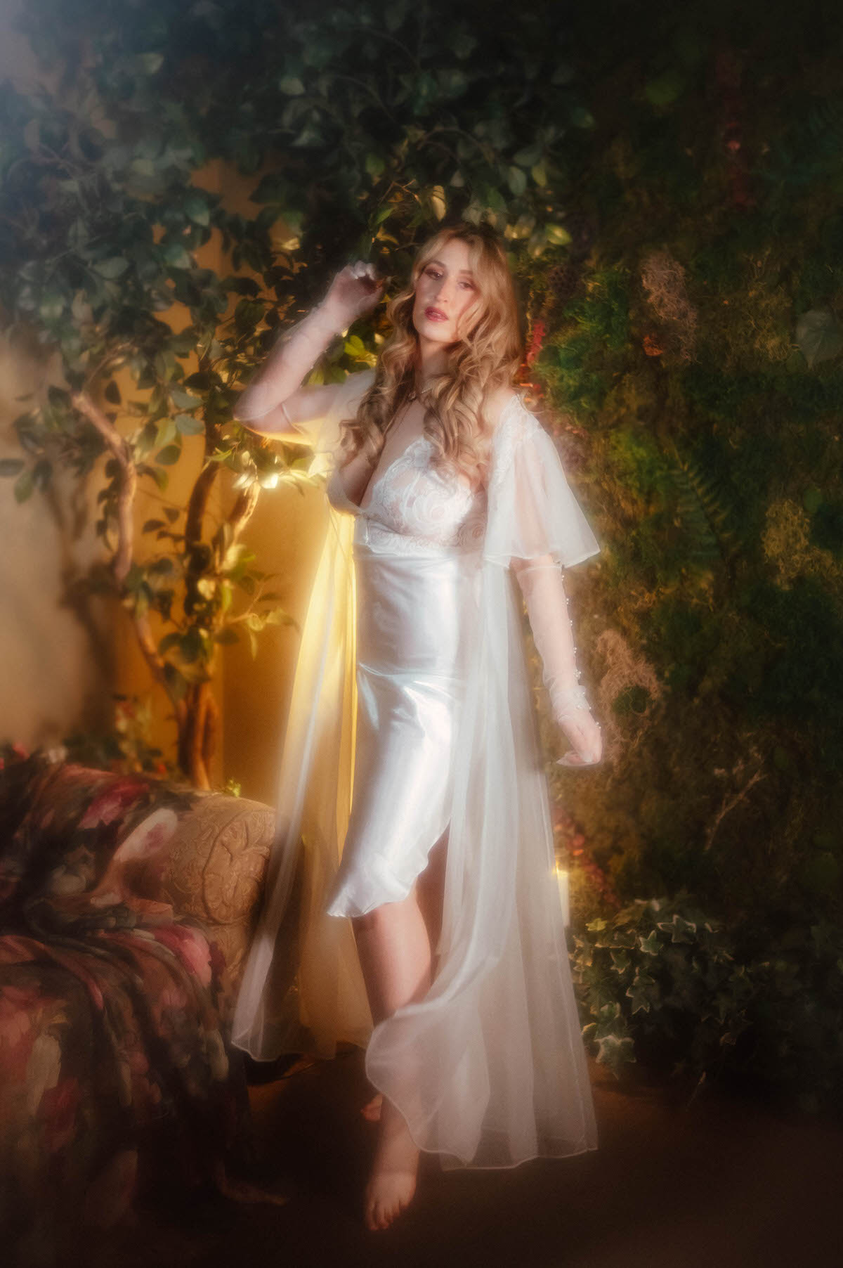 A woman in a vintage ivory satin slip dress with a lace bodice and vintage sheer chiffon robe reaches up into hanging ivy leaves, her pearl beaded glove catching warm golden light against a dark green backdrop, embodying the whimsical boudoir portraiture Dallas style of this dreamy boudoir photoshoot with Royal Lune Photo.