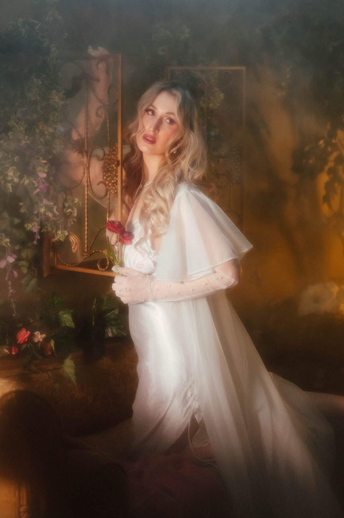 A blonde woman in an ivory satin slip dress with a sheer cape and pearl beaded gloves holds a single dark pink flower near an antique gold mirror surrounded by ivy, showcasing vintage boudoir photography DFW with warm, hazy lighting by Royal Lune Photo.