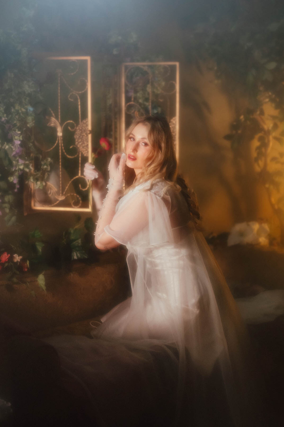 A woman holds a single dark pink flower near her chin with a pearl beaded glove, wearing a vintage sheer chiffon robe draped over one shoulder in front of gold framed mirrors and warm golden light, embodying the dreamy boudoir photoshoot style of Royal Lune Photo.