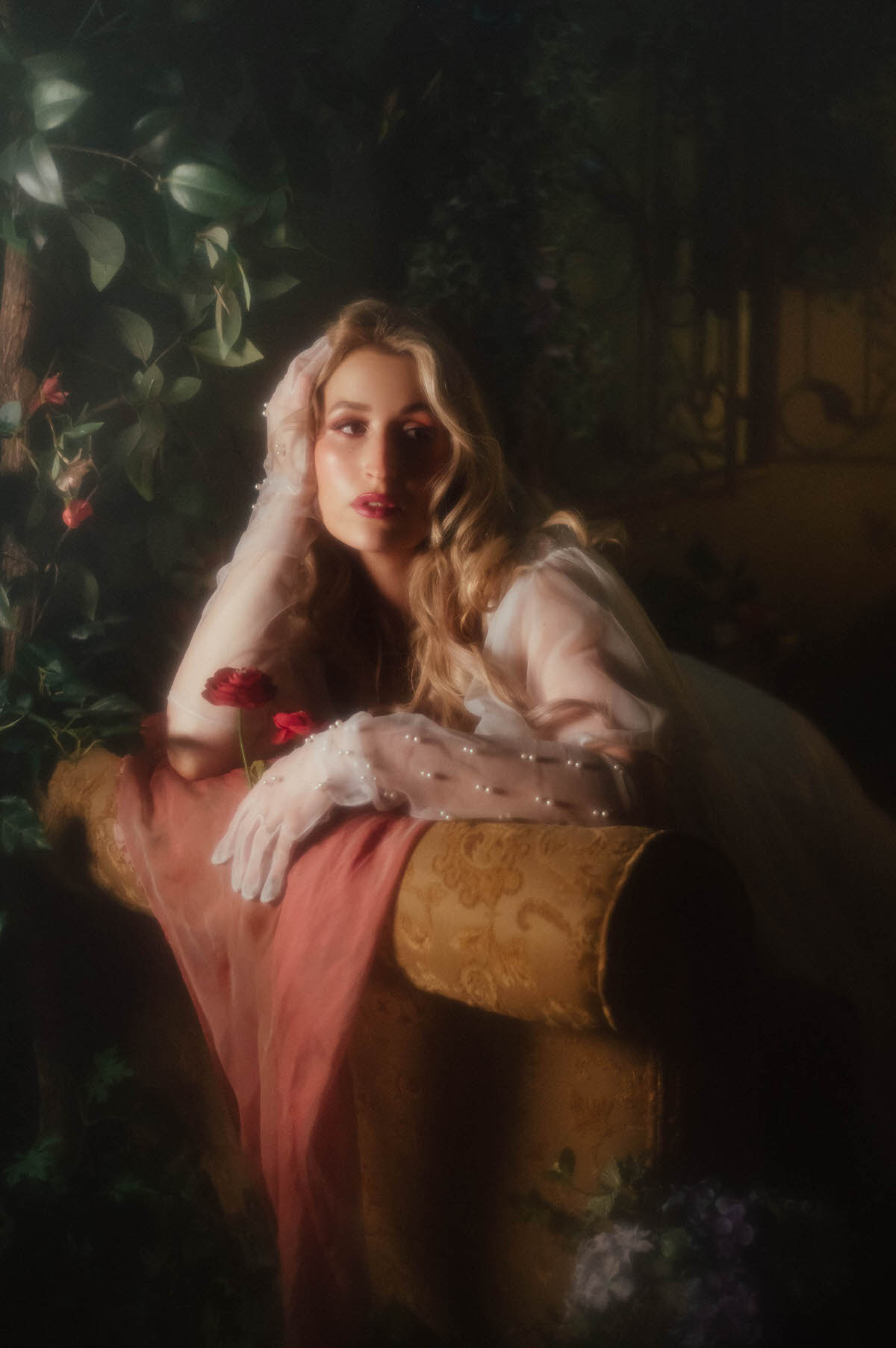 A woman leans over the arm of a gold vintage sofa draped in coral pink fabric, her vintage sheer chiffon robe falling over her head like a veil as she holds dark pink florals in her pearl beaded glove, adding a vintage boudoir photography DFW mood to this dreamy boudoir photoshoot with Royal Lune Photo.