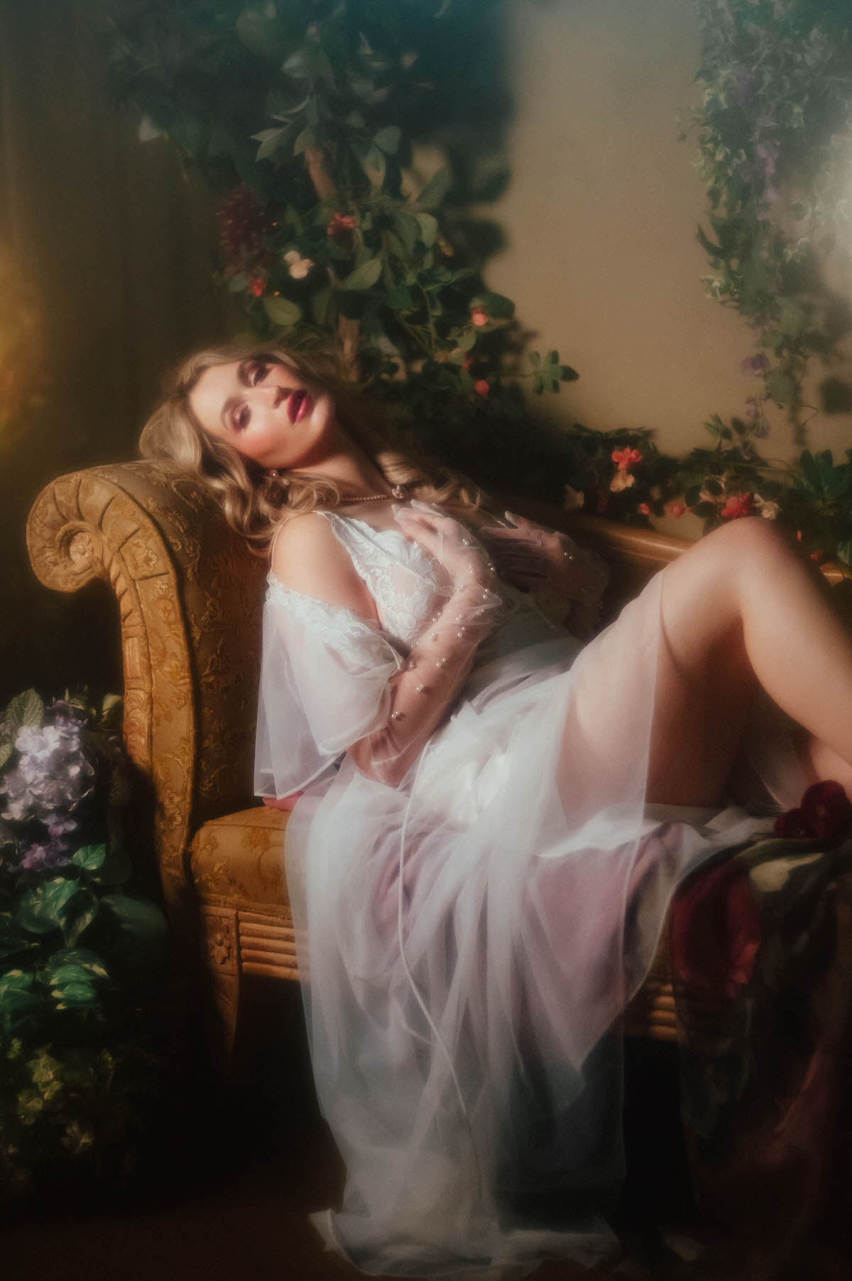 A woman reclines on an ornate gold chaise with her head tilted back and eyes closed, wearing a vintage ivory satin slip and vintage sheer chiffon robe with pearl beaded gloves against a backdrop of pink and red blooms, capturing the soft light intimate portraiture of this dreamy boudoir photoshoot with Royal Lune Photo.