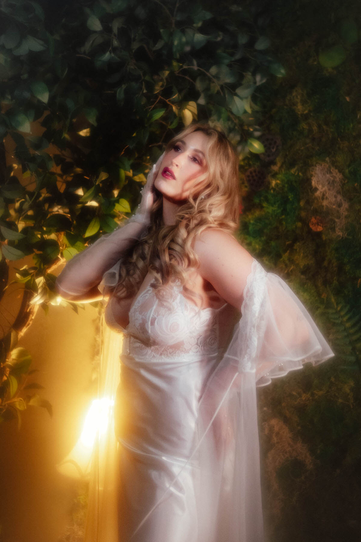 A woman in a vintage ivory lace bodice and vintage sheer nylon robe tilts her head back with a pearl beaded gloved hand near her face, warm golden light glowing behind her against a backdrop of ivy leaves, capturing soft light intimate portraiture for this dreamy boudoir photoshoot with Royal Lune Photo.