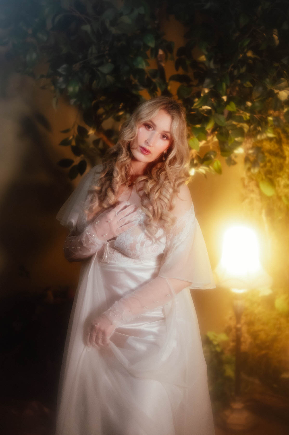 A woman in a vintage ivory satin slip and vintage sheer chiffon robe rests a pearl beaded gloved hand on her chest while gazing softly at the camera beside a glowing lamp and trailing greenery, showcasing the fine art boudoir photography Dallas style of Royal Lune Photo.
