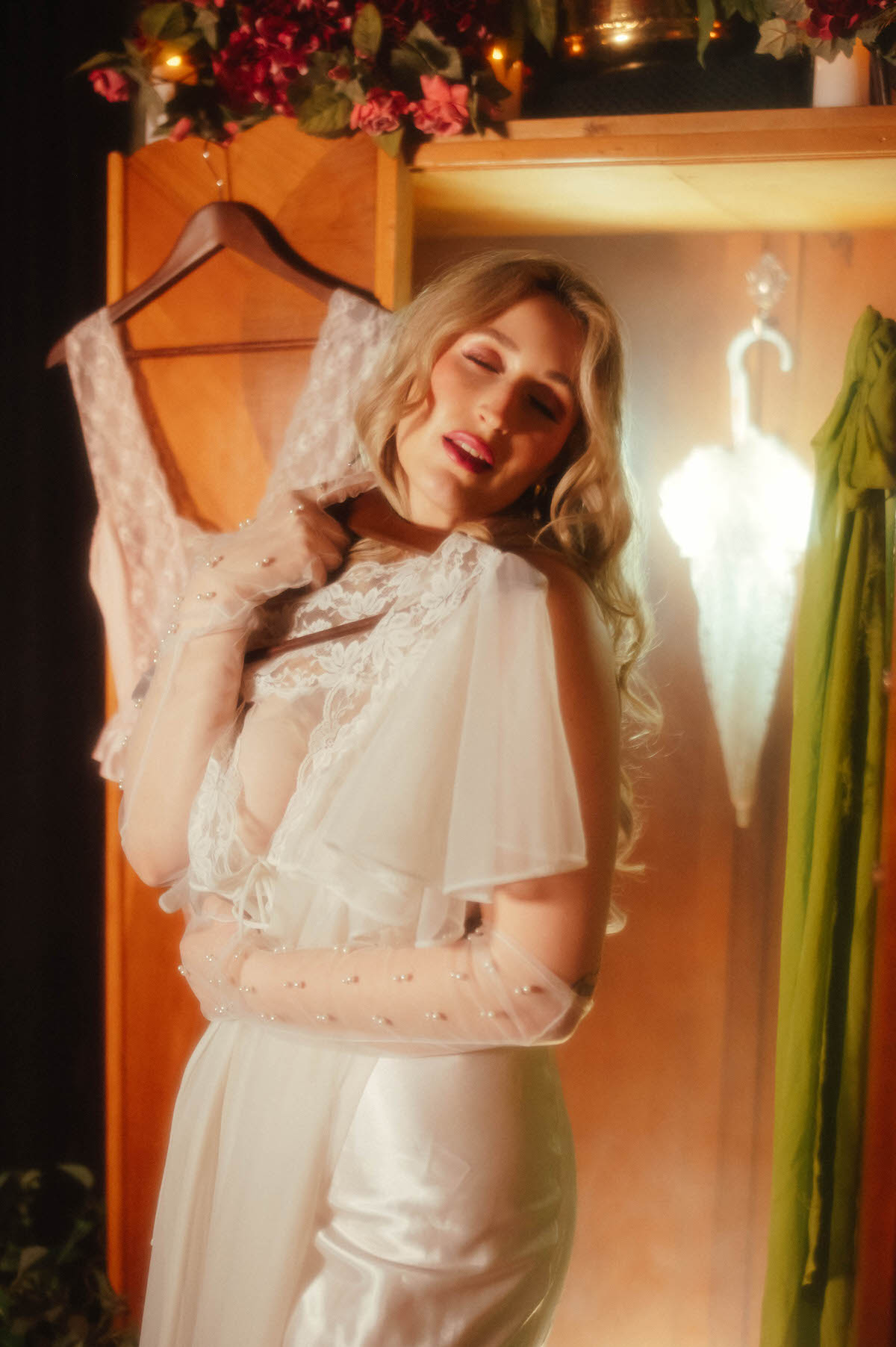 A close up shows a woman tilting her head back with her eyes closed while holding a vintage pink lace teddy near her shoulder, wearing a vintage ivory lace bodice and pearl beaded gloves in front of a vintage art deco armoire, part of the soft light intimate portraiture in this dreamy boudoir photoshoot with Royal Lune Photo.